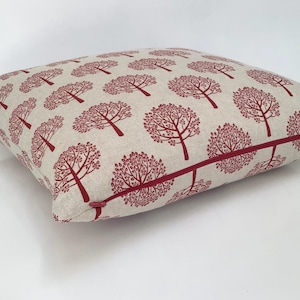 May include: Square cushion featuring a repeating pattern of red trees on a beige background. The cushion has a red zip and trim, and is likely made of a linen or cotton blend fabric. Suitable for home decor.