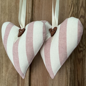 May include: Two heart-shaped fabric decorations with vertical stripes in white and pale red. Each heart has a small wooden button and a cream ribbon for hanging. They are displayed against a wooden background.