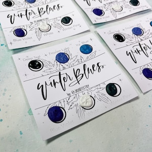 May include: Cards displaying the text "Winter Blues" and colour samples. Each card shows three colours: Blue Violet, White, and Winter Pine, alongside Deep Teal, Frozen Lake, and Icy Blue. The cards are set against a light blue and white watercolour background.