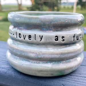 May include: A cylindrical ceramic mug with a glossy, multi-toned light blue and grey glaze. The exterior features three horizontal ridges and the stamped black text "...lovely as fuck" encircling the middle section of the vessel.