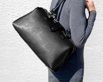 May include: A black rectangular shoulder bag with a smooth finish, featuring a top handle, a secure flap closure with a snap button, and silver-toned hardware attachments for the straps, worn over the shoulder against a grey long-sleeved top.