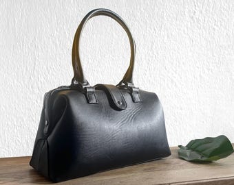 May include: A black handbag with a structured doctor bag silhouette, featuring a single rounded top handle, a central snap-button closure tab, and a zippered top opening. The bag has a smooth, matte finish and a wide, flat base design.