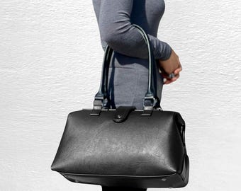 May include: A black structured handbag with a smooth finish, featuring dual top handles, a central snap-button closure, and a rectangular base with visible metal feet. The bag has a clean, minimalist design with dark hardware accents.