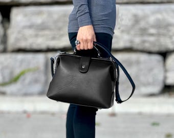 May include: A structured black handbag with a smooth finish, featuring a top handle, a secure flap closure with a central snap button, and a detachable shoulder strap. The bag has a classic doctor-style frame silhouette with clean lines.