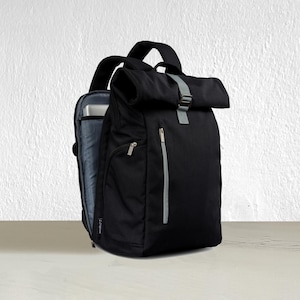May include: Black backpack with a roll-top closure and a gray strap. The backpack has a zippered front pocket and a side pocket. The interior is visible, revealing a laptop sleeve. The bag has a top handle and adjustable shoulder straps.