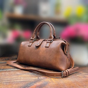 May include: A brown leather satchel bag with a top handle and a detachable shoulder strap. The bag has a rectangular shape with a curved top and metal hardware. The bag is sitting on a wooden surface.
