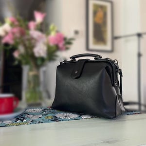 May include: A black doctor-style handbag with a structured frame, a top handle, and a central flap closure with a snap button. The bag features a smooth, matte finish and metal hardware attachments on the sides for a shoulder strap.