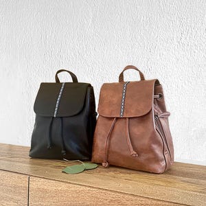 May include: Two leather backpacks, one black and one brown, sit side-by-side on a wooden surface. Each backpack has a flap closure with a decorative stitched stripe down the center. The brown backpack has a drawstring closure and a side zipper.