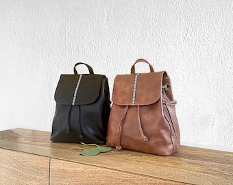 May include: Two leather backpacks, one black and one brown, sit side-by-side on a wooden surface. Each backpack has a flap closure with a decorative stitched stripe down the center. The brown backpack has a drawstring closure and a side zipper.