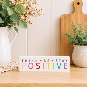May include: A rectangular white wooden block sign featuring the black text THINK BE STAY and the word POSITIVE in large, multicolored letters. Two small pink hearts are positioned between the words on the top line of the sign.