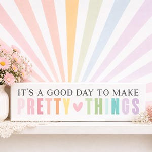 May include: A rectangular white wooden sign featuring the black text IT'S A GOOD DAY TO MAKE and the colorful text PRETTY THINGS with a small pink heart in the center. The sign is displayed alongside a vase of pink daisies and white flowers.