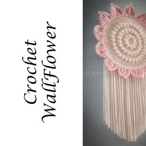 May include: A pink and cream crochet wall flower with a fringe of white yarn. The flower is made with a circular design and has a textured appearance.