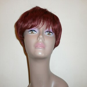 May include: A mannequin head displaying a short, burgundy wig. The wig is styled with layers and a side parting, offering a natural appearance. The mannequin features painted facial details, including large eyes and full lips.