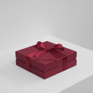 May include: A square, deep burgundy gift box with a matching satin ribbon tied in a bow on top. The box features a two-piece construction with a lid and base, and the word BLONDE is printed in small gold letters on the side of the lower section.