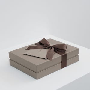 Könnte beinhalten: Eine rechteckige, taupefarbene Geschenkbox mit zweiteiligem Deckel. Die Box ist mit einem dunkelbraunen Satinband umwickelt, das oben zu einer Schleife gebunden ist, und eine passende taupefarbene Karte steckt ordentlich unter dem Band.