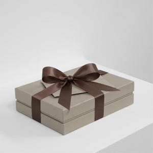 Könnte beinhalten: Eine rechteckige Geschenkbox in hellem Taupe mit einer glatten, matten Oberfläche. Die Box ist mit einem dunkelbraunen Satinband umwickelt, das zu einer großen, mittigen Schleife auf dem Deckel gebunden ist und eine klare, strukturierte Optik bietet.