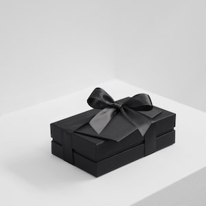 Puede incluir: Una caja de regalo rectangular de color negro con acabado mate, envuelta con una cinta de satén negra a juego atada en un lazo en la parte superior central. La caja presenta una estructura de dos piezas con tapa y base, de aspecto uniforme.