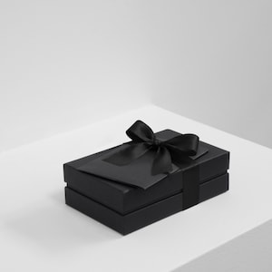 Könnte beinhalten: Eine rechteckige schwarze Geschenkbox mit matter Oberfläche, die mit einer passenden schwarzen Satinschleife auf dem Deckel verziert ist. Die Box besteht aus einem zweiteiligen Design mit Unterteil und abnehmbarem Deckel und wirkt einheitlich.