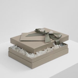 Può includere: Una scatola regalo rettangolare color beige con coperchio, legata con un nastro di raso verde salvia e decorata con piccoli fiori secchi bianchi. La scatola è socchiusa, rivelando uno strato di materiale di riempimento bianco e una busta beige.
