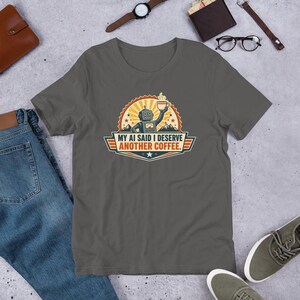 Puede incluir: Una camiseta gris carbón con un gráfico de un robot de estilo retro sosteniendo una taza de café humeante. El diseño incluye un fondo de rayos de sol, siluetas de montañas y el texto "MY AI SAID I DESERVE ANOTHER COFFEE." en letras negritas.