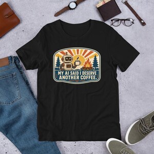 Puede incluir: Una camiseta negra de manga corta con un gráfico de un robot sosteniendo una taza de café junto a una fogata, un sol y pinos. El texto dice "MY AI SAID I DESERVE ANOTHER COFFEE." El diseño utiliza una paleta de colores retro en naranja, amarillo y azul.
