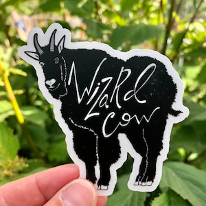 May include: A black and white sticker featuring a stylized illustration of a goat with the text "Wizard Cow" in white script. The sticker is held against a backdrop of green foliage.