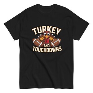 May include: Black t-shirt with a cartoon turkey design. The design includes the words "TURKEY and TOUCHDOWNS" in white and gold. The turkey has a red wattle and yellow beak, with brown footballs on either side.