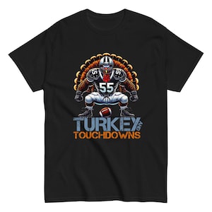 Könnte beinhalten: Schwarzes T-Shirt mit einer Cartoon-Truthahnfigur in Football-Ausrüstung, Nummer 55 auf dem Trikot. Der Truthahn hockt und hält einen Football, mit dem Text "TURKEY AND TOUCHDOWNS" in Blau und Orange.