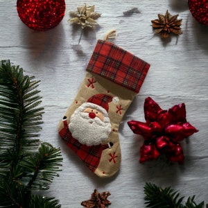 Op de afbeelding: Een beige kerstsok met een rood-zwarte geruite manchet. De sok is versierd met een geborduurde kerstman met een witte baard en een rode muts. De sok is versierd met rode en witte sneeuwvlokken.