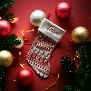 以下が含まれることがあります： 赤い白のクリスマスストッキング。白いカフスには赤い刺繍で「Theo」と書いてあります。ストッキングは、金と白のオーナメントと松の枝で囲まれています。