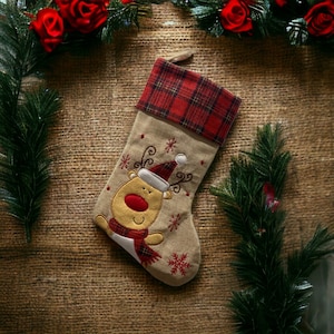 Peut inclure: Une chaussette de Noël marron avec un revers en tartan rouge et noir. La chaussette est ornée d'un renne brodé avec un nez rouge et une écharpe rouge et blanche.