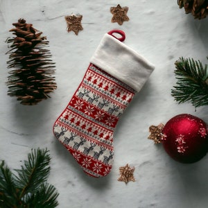 Può includere: Un calzino di Natale rosso e bianco con un polsino bianco e un motivo a renne. Il calzino è su una superficie di marmo bianco con rami di pino e ornamenti.