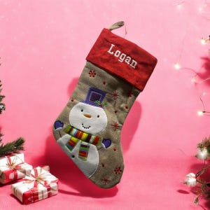 Può includere: Un calzino di Natale marrone e beige con un disegno di pupazzo di neve. Il calzino ha un polsino rosso con il nome "Logan" ricamato.