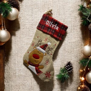 Könnte beinhalten: Ein beiger Weihnachtssocken mit einem roten und schwarzen karierten Bündchen. Der Socken ist mit einem gestickten Rentier mit roter Nase und einer roten und weißen Weihnachtsmütze verziert. Der Name "Olivia" ist auf das Bündchen gestickt.