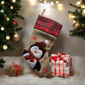Op de afbeelding: Een kerstsok met een pinguïnontwerp. De sok is beige met rode sneeuwvlokaccenten en een rode geruite manchet. Een pinguïn met een hoed en sjaal staat op de voorkant. Cadeaus verpakt in wit papier met rode linten staan op de achtergrond.