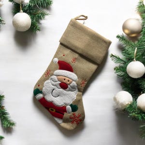 Può includere: Un calzino di Natale in tela di juta beige con un'applicazione di Babbo Natale. Il calzino ha un polsino bianco ed è decorato con fiocchi di neve.