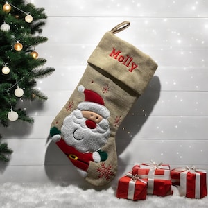 Op de afbeelding: Een beige jute kerstsok met een Santa Claus applicatie en de naam "Molly" geborduurd op de manchet. De sok is gevuld met rood ingepakte cadeaus.