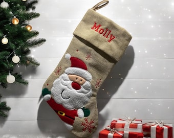 May include: A beige burlap Christmas stocking with a Santa Claus applique and the name "Molly" embroidered on the cuff. The stocking is filled with red wrapped gifts.