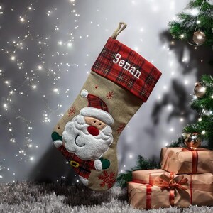 以下が含まれることがあります： 赤と黒のタータンチェックの袖口が付いた、名前入りの麻のクリスマスストッキング。ストッキングには、白いひげと赤い帽子をかぶったサンタクロースのフェルトアップリケが付いています。ストッキングには「Sean」という名前が書かれています。