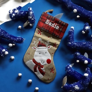 May include: A festive Christmas stocking featuring a reindeer design. The stocking is tan with a red plaid cuff, embroidered with the name "Sadie". The reindeer has a red nose and scarf. The background is blue with decorative elements.