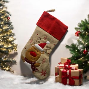 Può includere: Un calzino di Natale marrone e beige con un polsino in feltro rosso. Il calzino presenta una renna ricamata con un naso rosso e un cappello di Babbo Natale rosso e bianco. Il calzino è decorato con fiocchi di neve.