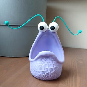 Könnte beinhalten: Ein lila und türkisfarbenes Spielzeug aus Plastik mit großen Augen und einem weit geöffneten Mund. Das Spielzeug hat eine strukturierte Oberfläche und ist in Form einer Kreatur mit zwei Antennen.