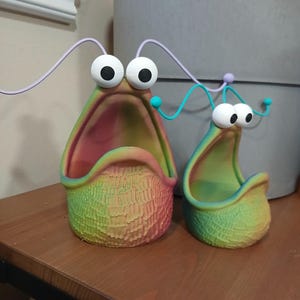 May include: Two colorful, 3D printed planters shaped like a cartoon monster with a wide open mouth and large eyes. The planters are made of a textured material and have a rainbow gradient color scheme.