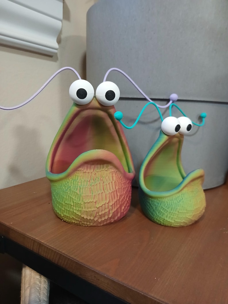 May include: Two colorful, 3D printed planters shaped like a cartoon monster with a wide open mouth and large eyes. The planters are made of a textured material and have a rainbow gradient color scheme.