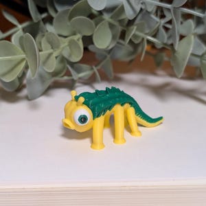 Puede incluir: Una criatura de juguete amarilla y verde con un ojo grande y una espalda puntiaguda. El juguete tiene cuatro patas amarillas y una cola verde. La criatura está sobre un fondo neutro con follaje verde.