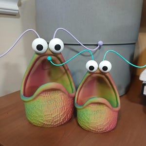 May include: Two colorful, ceramic planters shaped like open mouths with large eyes and a textured surface. The planters are a rainbow of colors, including red, orange, yellow, green, blue, and purple.