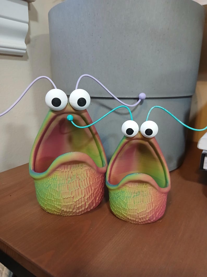 May include: Two colorful, ceramic planters shaped like open mouths with large eyes and a textured surface. The planters are a rainbow of colors, including red, orange, yellow, green, blue, and purple.