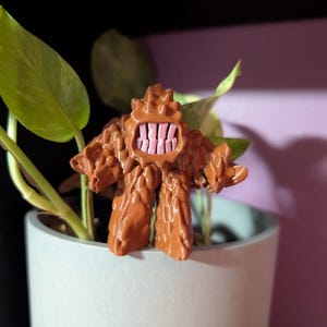 May include: A brown figurine of a creature with a textured surface and a wide, toothy grin. The figurine is positioned in front of a green plant in a white pot, creating a playful contrast of colours and textures.