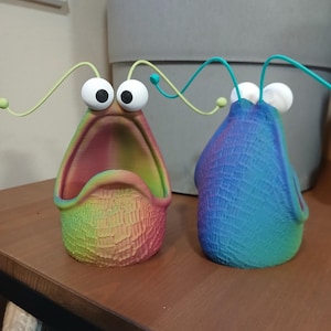 May include: Two colorful, 3D printed planters shaped like a creature with large eyes and a wide open mouth. The planters are made with a rainbow gradient color scheme.