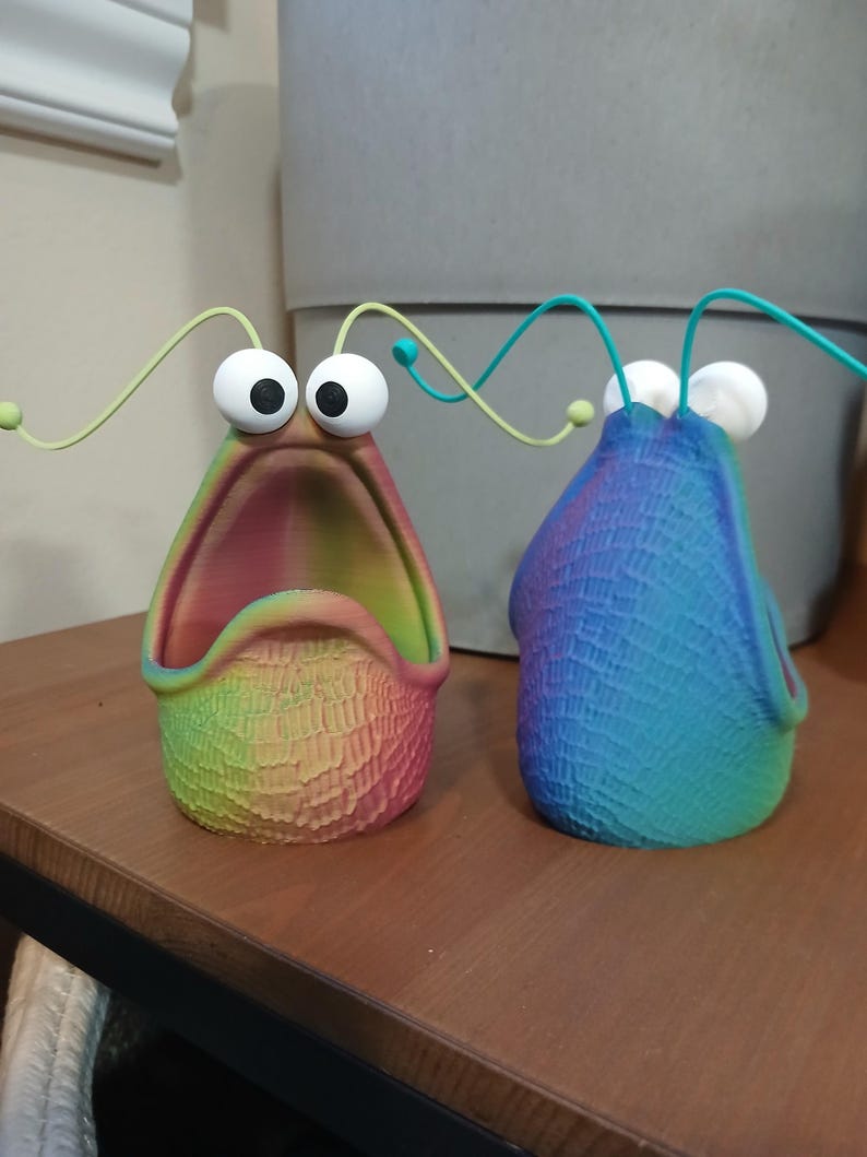 May include: Two colorful, 3D printed planters shaped like a creature with large eyes and a wide open mouth. The planters are made with a rainbow gradient color scheme.
